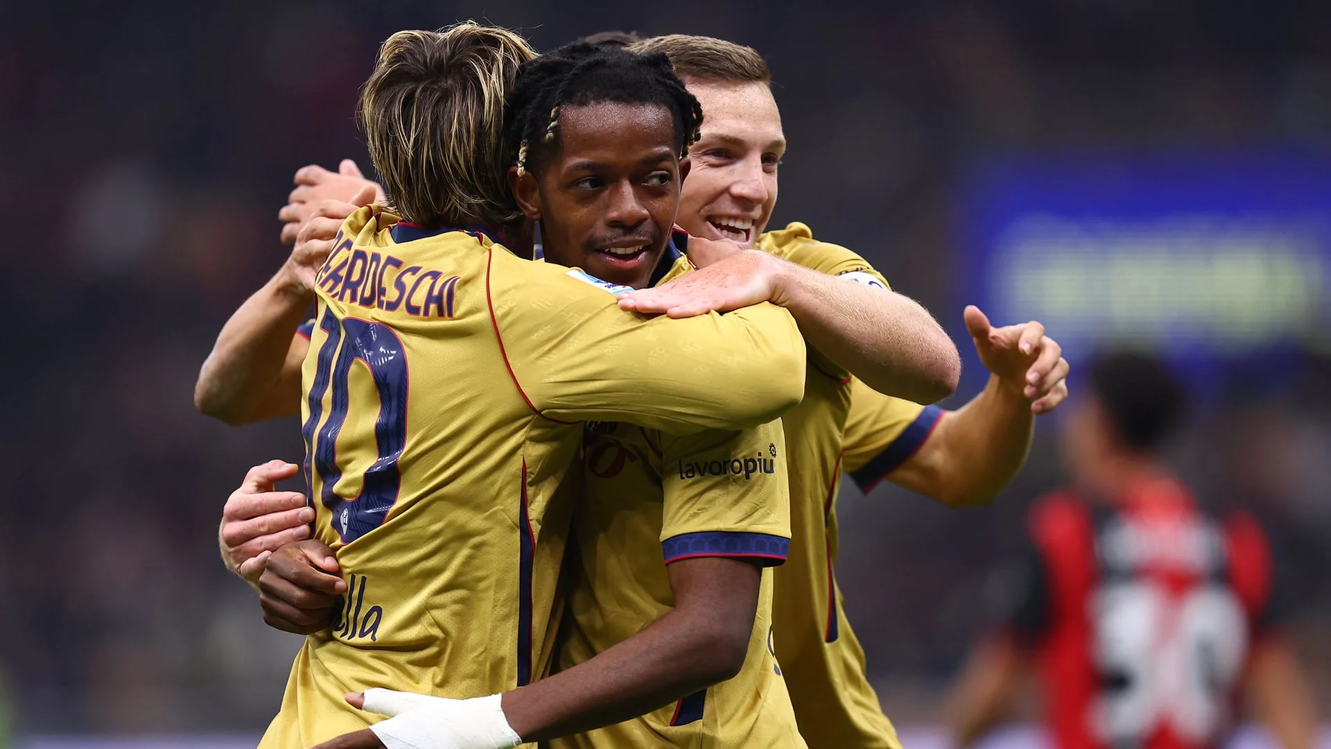 Bologna forward Samuel Mbangula, in the club's yellow away kit, smiles as team-mates including Federico Bernardeschi (No 10) embrace him after a goal at San Siro, with a blurred Milan player in red and black in the background.