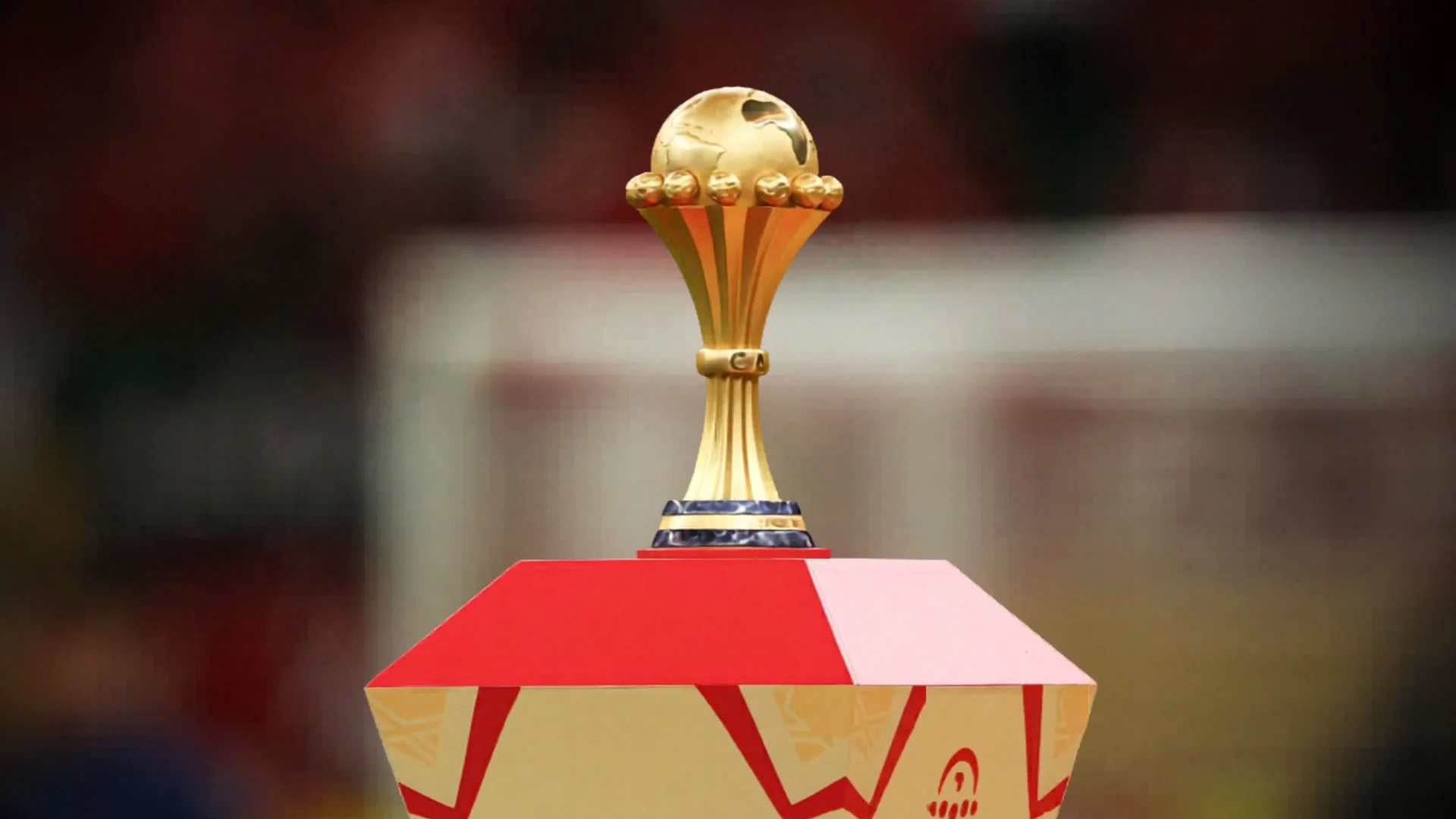 The Africa Cup of Nations trophy displayed on a red and gold plinth inside a stadium