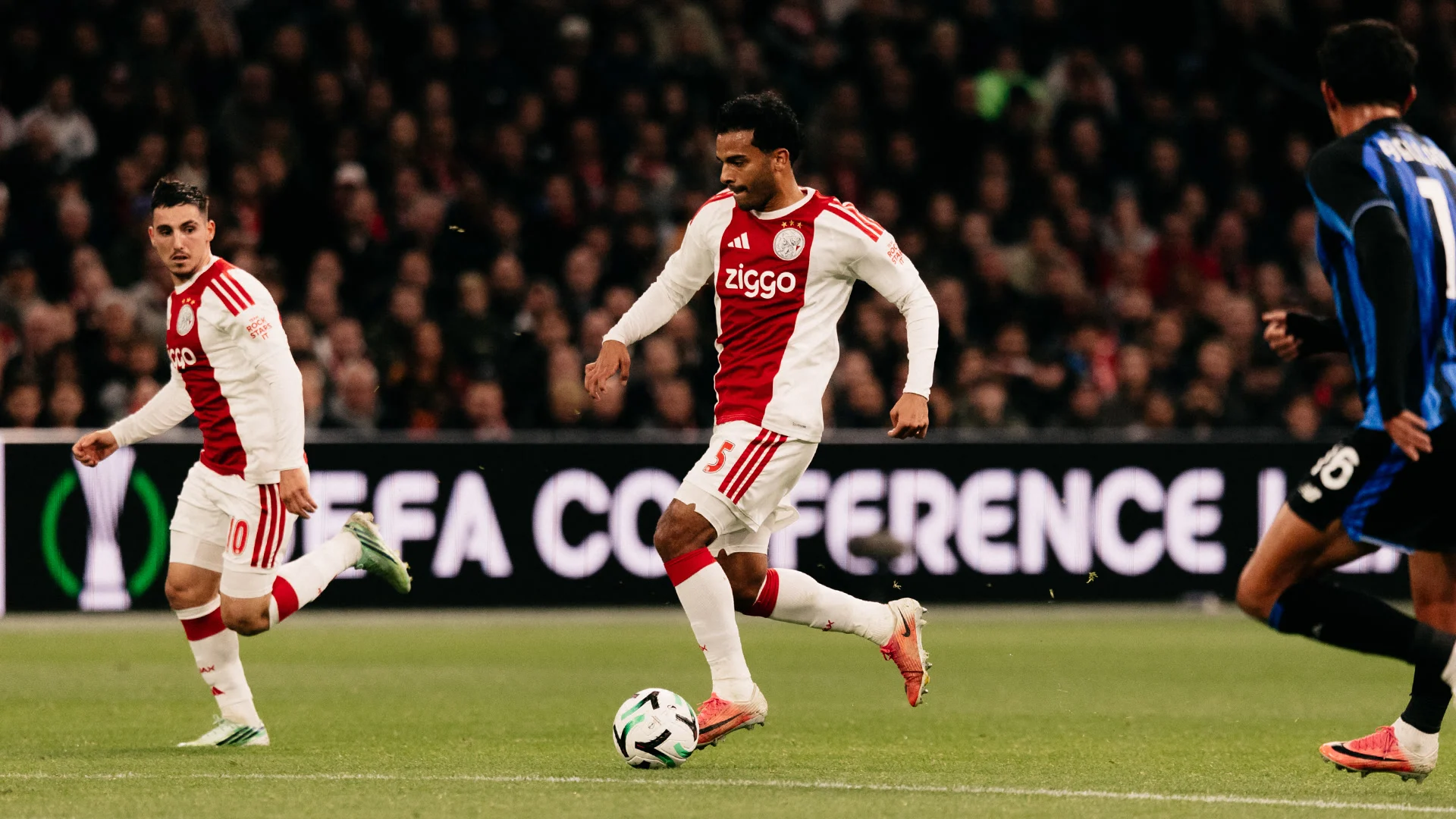 Ajax defender Owen Wijndal, wearing number 5 in Ajax's red and white home kit, runs with the ball as teammate Oscar Gloukh (number 10) follows and an Atalanta player in the blue and black kit closes in, at the Johan Cruijff ArenA. Photo: AFC Ajax Socials