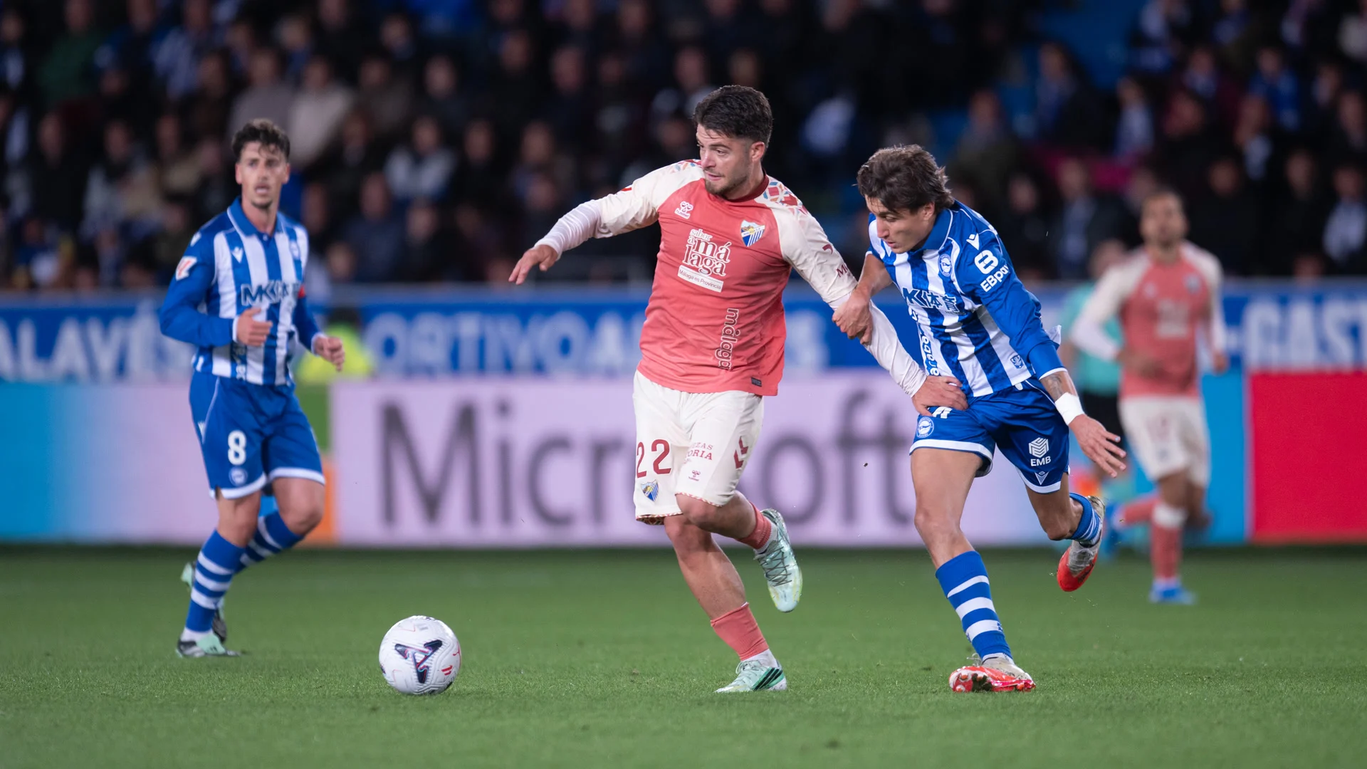 Málaga midfielder Dani Lorenzo, number 22 in the club's coral away shirt, holds off an Alavés player in a blue-and-white striped shirt during the La Liga match at Mendizorroza on 23 October 2026. Photo: Málaga CF