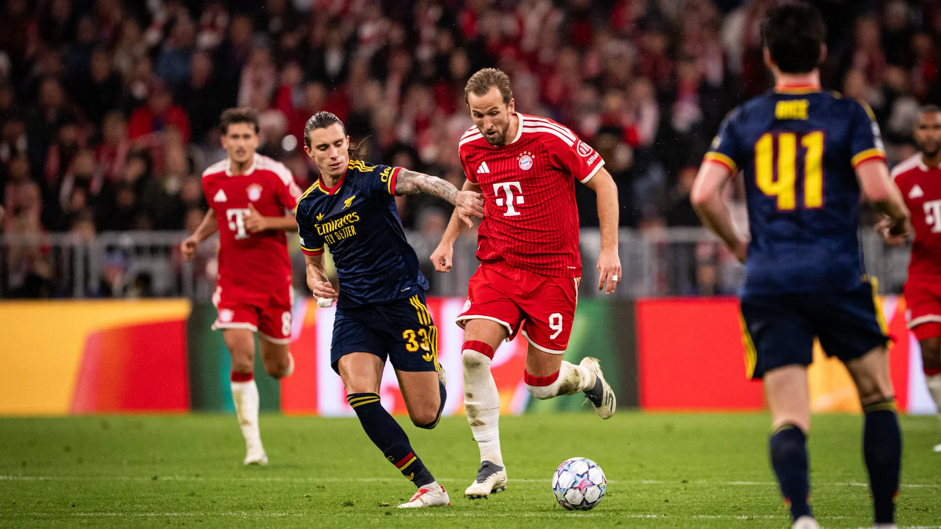 Harry Kane, in Bayern Munich's red home kit with the number 9, runs with the ball as Arsenal's Riccardo Calafiori (number 33, dark blue away kit) challenges him and Declan Rice (number 41) watches, at the Allianz Arena. Photo: F. Noever/FC Bayern via Getty Images