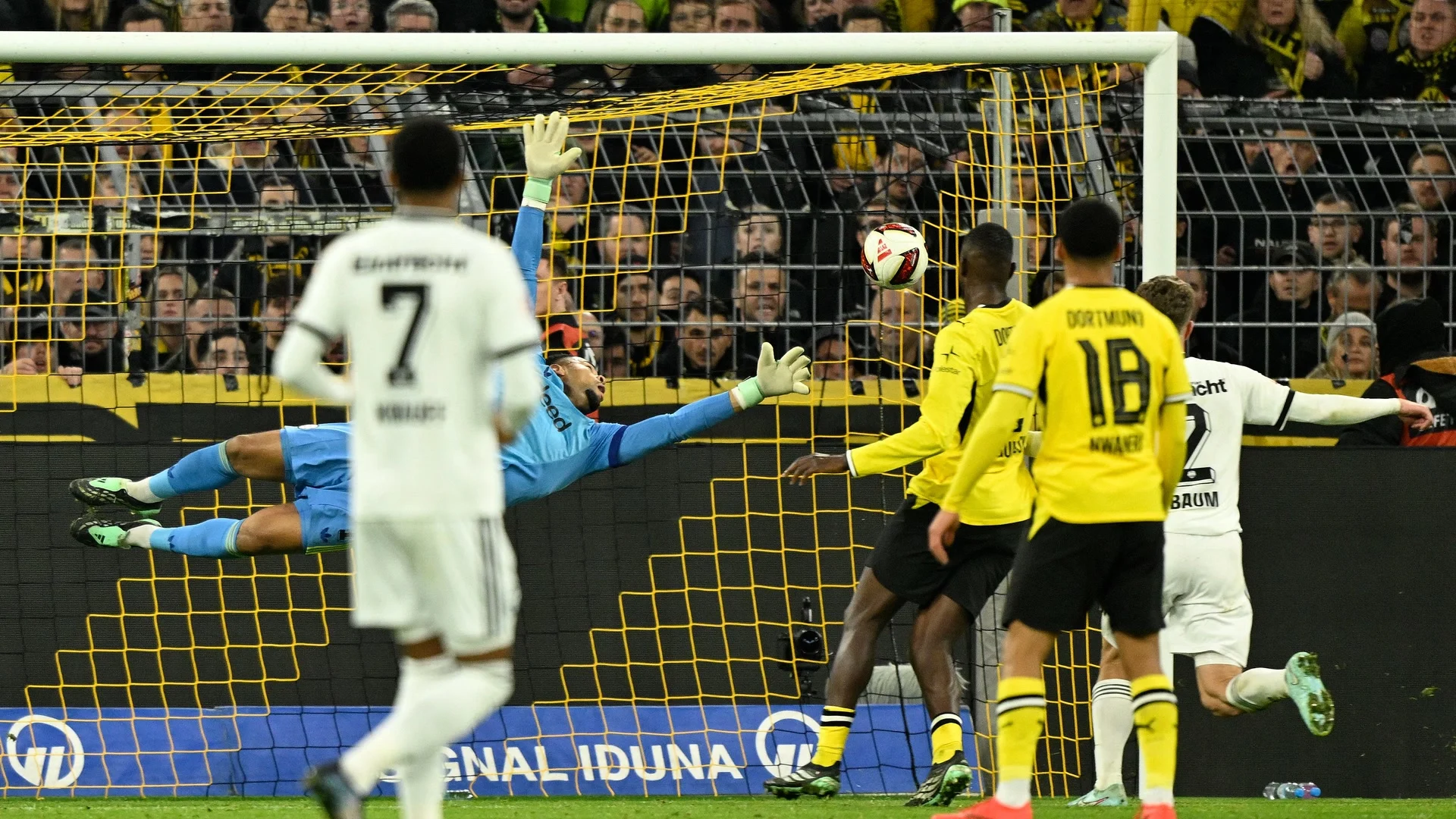 Eintracht Frankfurt goalkeeper Noah Atubolu, in light blue, dives at full stretch as the ball flies towards the top corner of his net, with Dortmund's Ethan Nwaneri (No 18) and Frankfurt's Elias Baum looking on at Signal Iduna Park on 24 October 2026. Photo: Ina Fassbender via Bundesliga.com