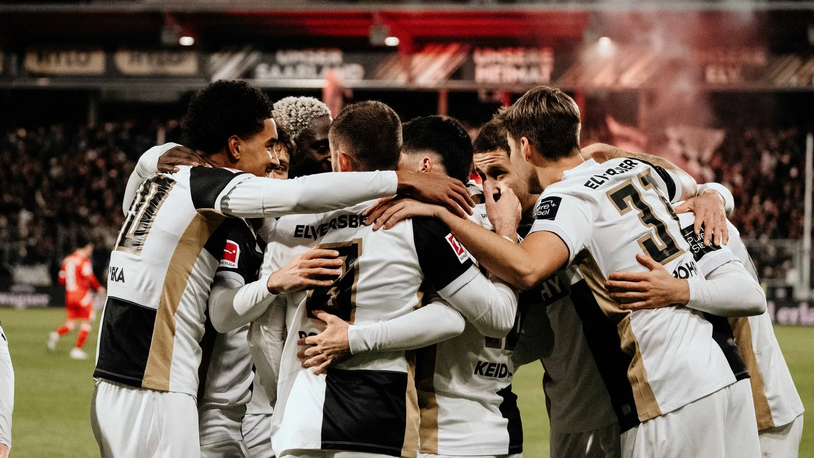 SV Elversberg players in white kits huddle together to celebrate a goal against Mainz at the Ursapharm-Arena an der Kaiserlinde