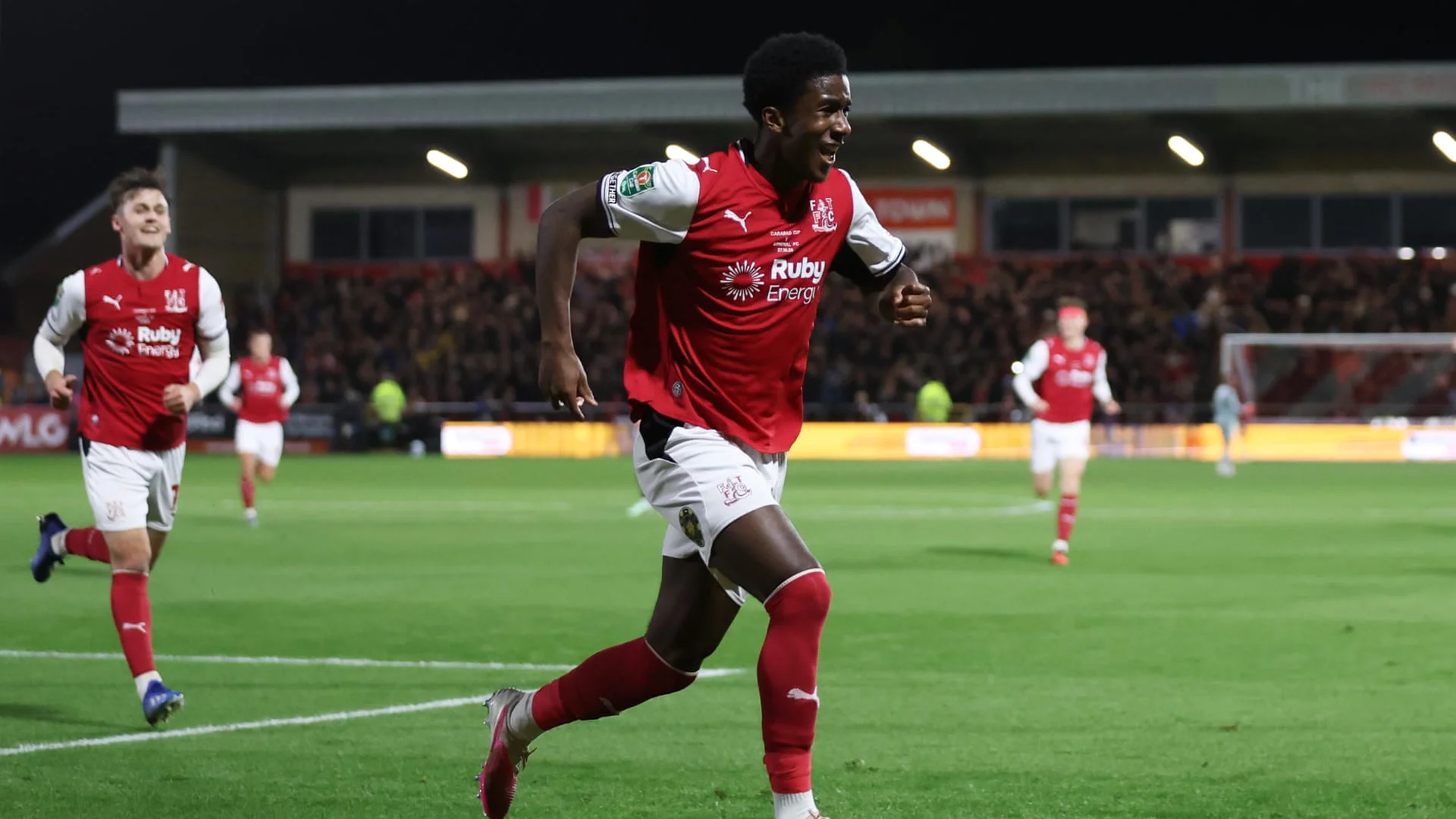 Fleetwood Town forward Aaron Loupalo-Bi, in the club's red and white home kit, runs away smiling after scoring against Arsenal at Highbury Stadium, with Fleetwood team-mates following behind.