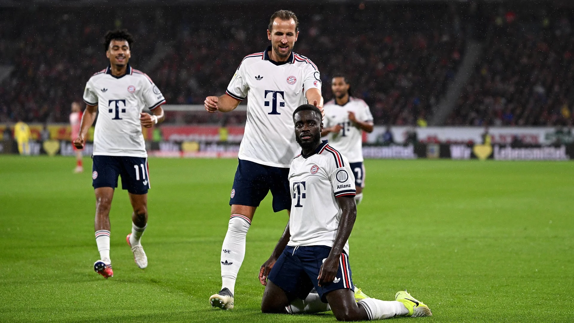 Bayern Munich defender Dayot Upamecano, in Bayern's white away shirt, kneels on the pitch celebrating his opening goal against SC Freiburg at the Europa-Park Stadion on 25 October 2026, with Harry Kane standing behind him and team-mates running in to join them. Photo: IMAGO/Pressefoto ULMER/Markus Ulmer via bundesliga.com