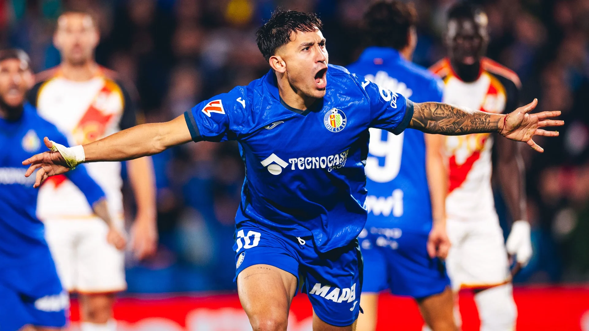 Getafe striker Martín Satriano (No 10) celebrates with his arms outstretched in Getafe's blue home kit during the 1-1 La Liga draw with Rayo Vallecano at the Coliseum, with Rayo players in white and red behind him. Photo: Getafe CF