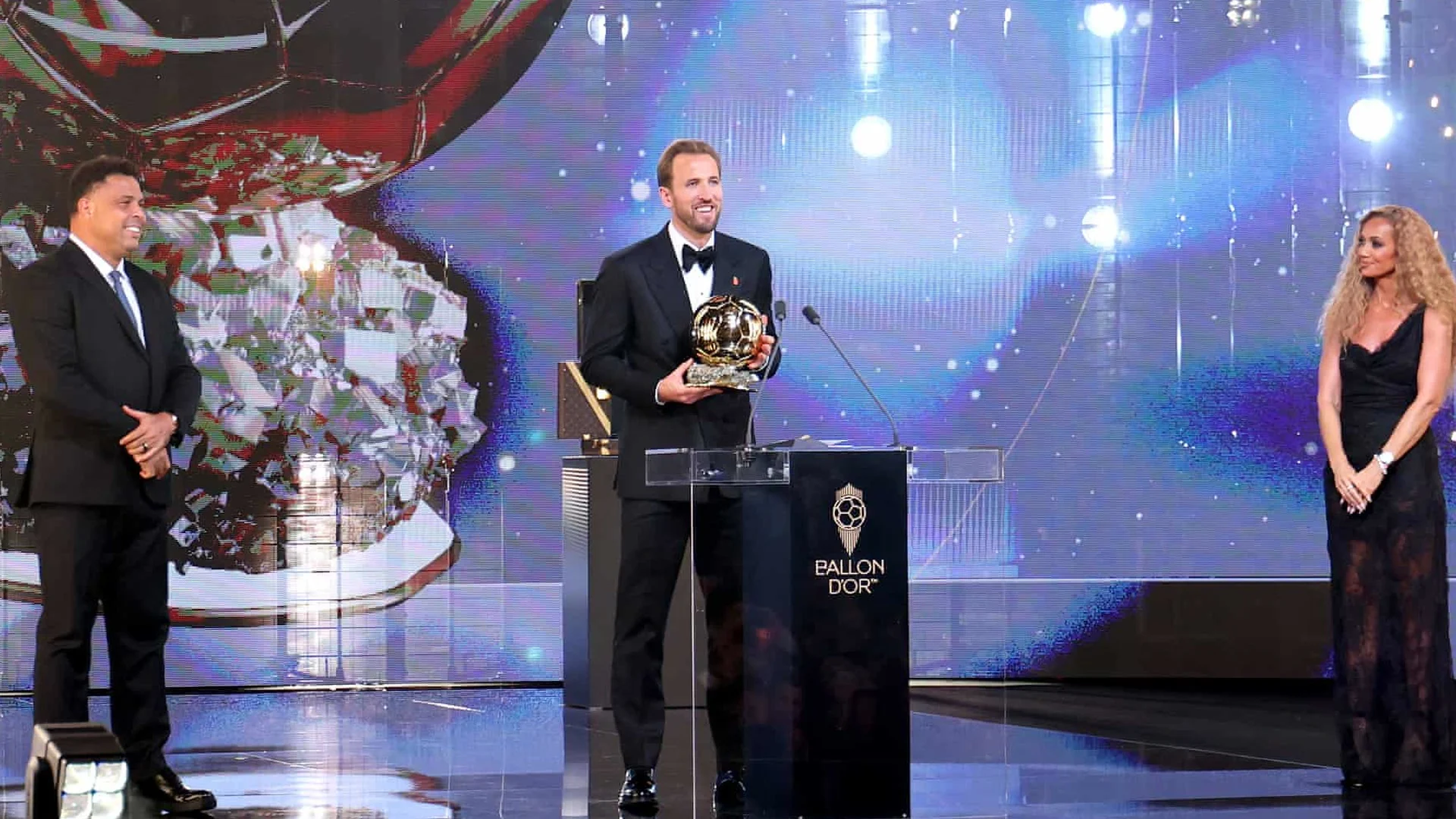 Harry Kane, in a black tuxedo and bow tie, holds the Ballon d'Or trophy at the lectern on stage at the London Palladium as Ronaldo Nazário and a ceremony host look on, 26 October 2026