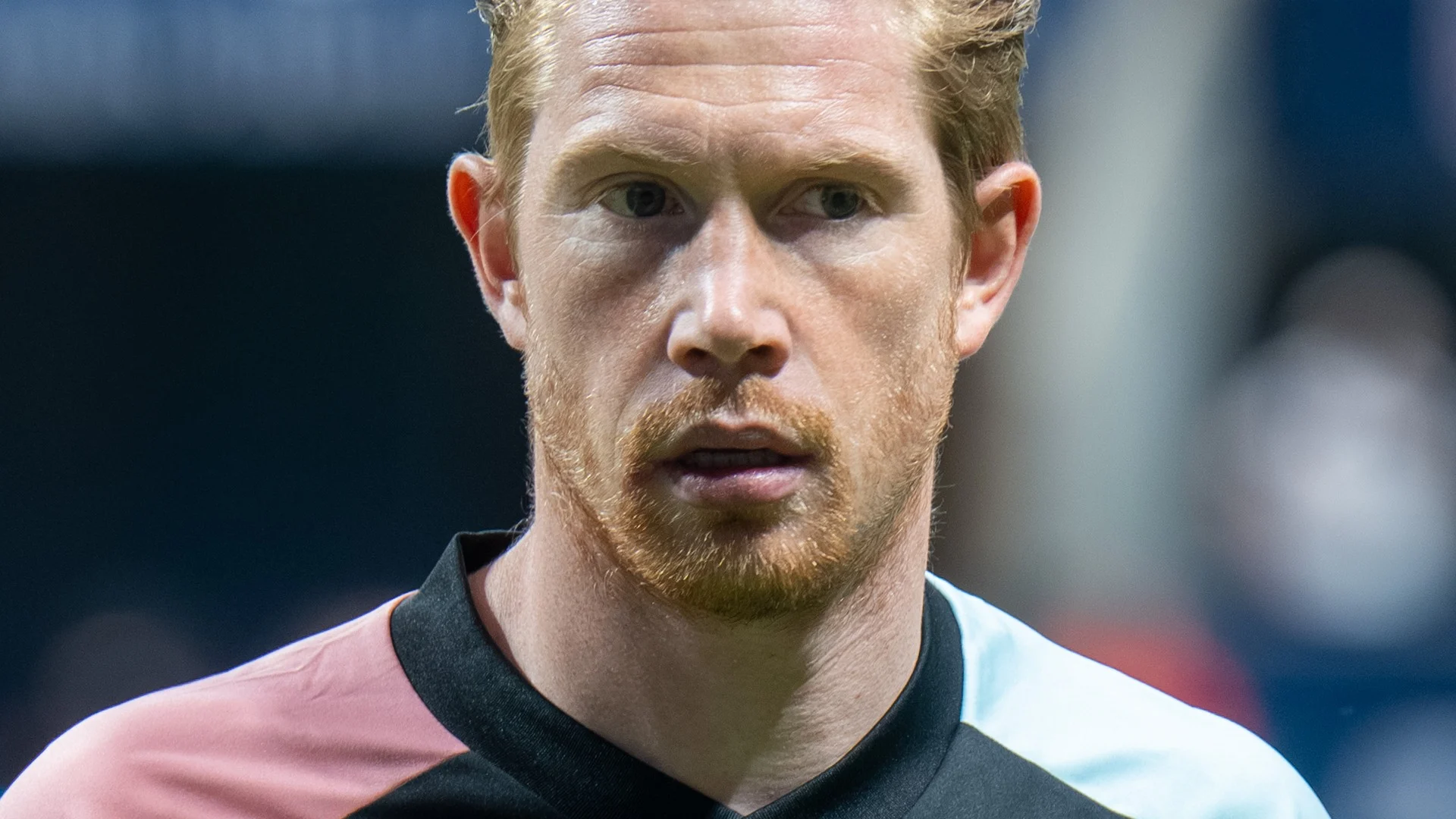 Kevin De Bruyne in a black Belgium training jersey with pink and light-blue sleeves looking focused in a stadium.