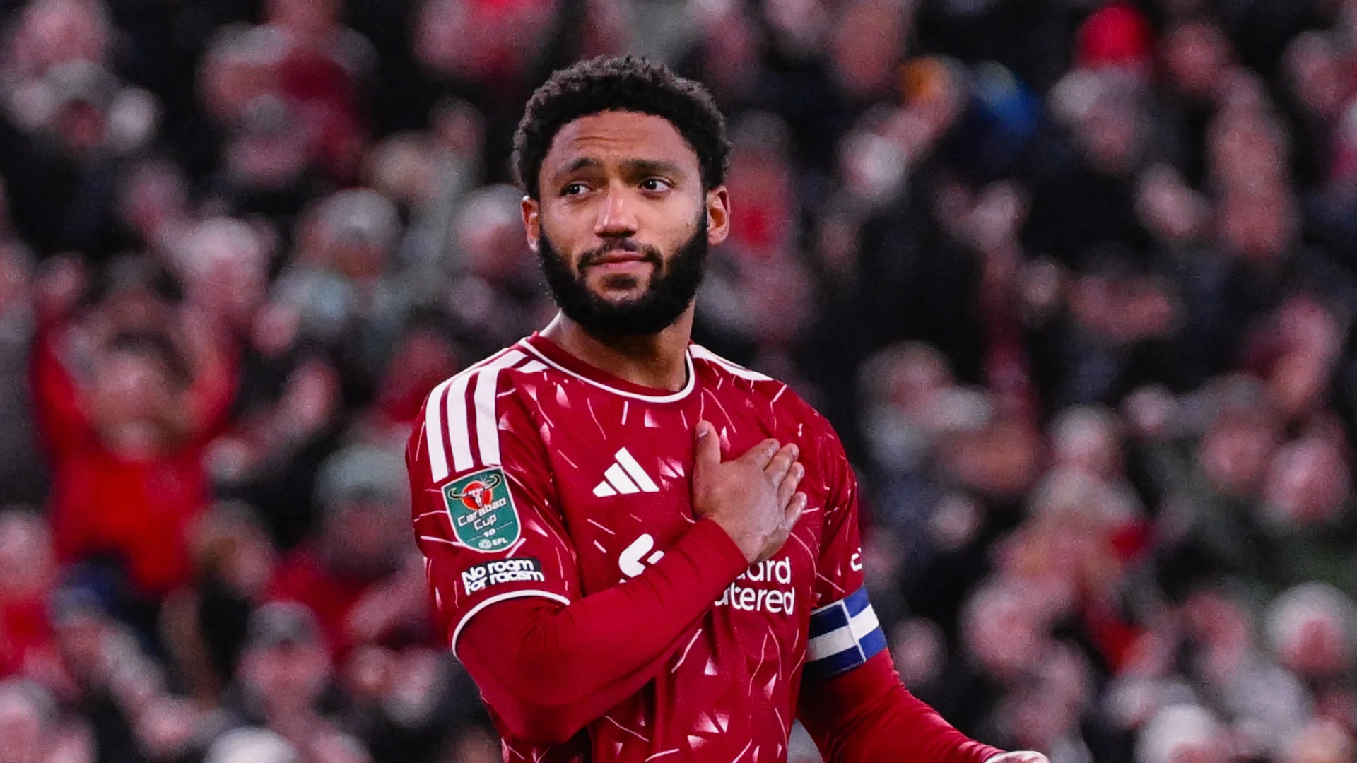 Liverpool defender Joe Gomez, wearing the captain's armband and a Carabao Cup sleeve badge, holds his hand to his chest in front of the Anfield crowd.
