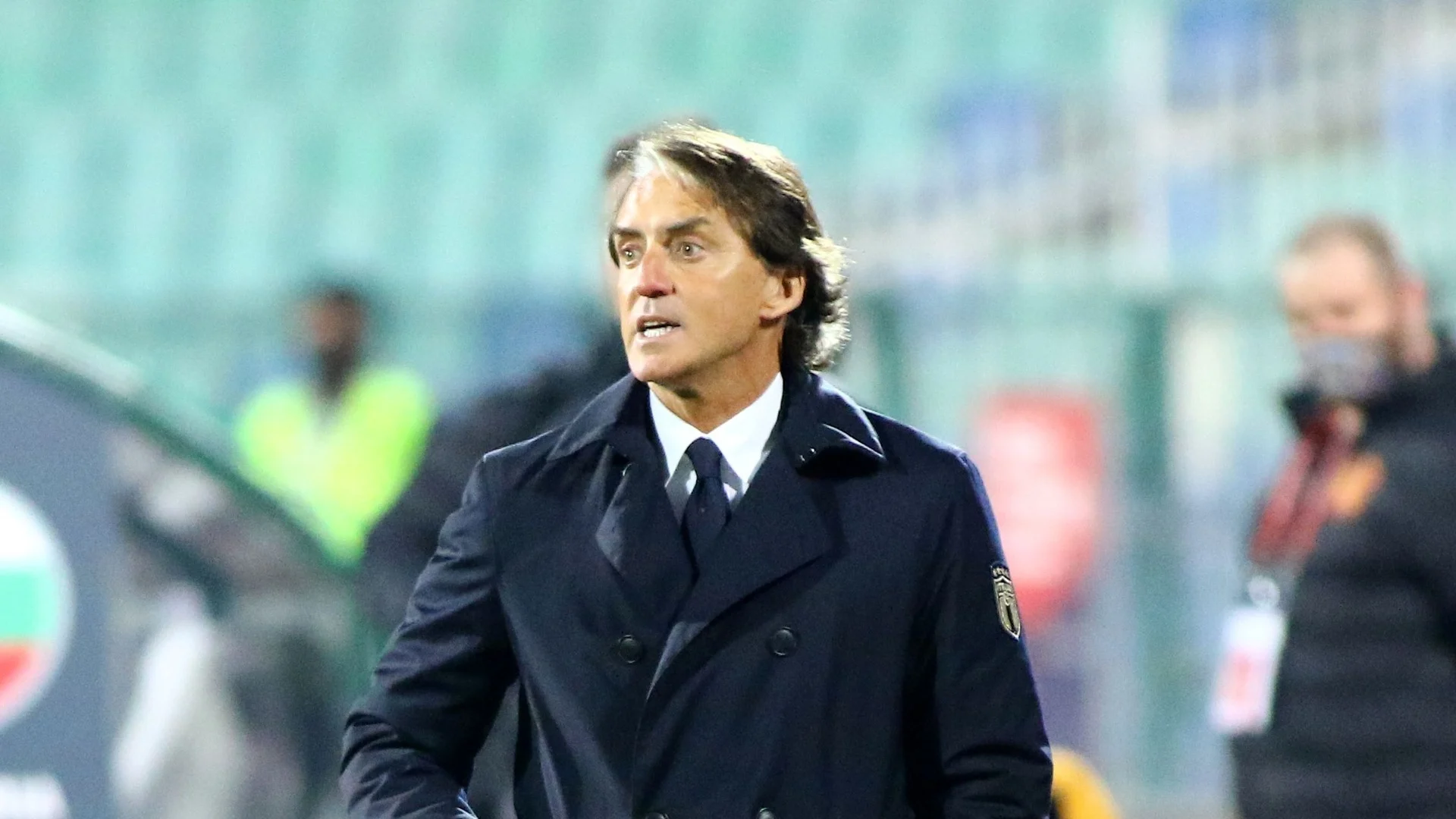 Roberto Mancini on the Italy touchline in a dark FIGC-crested coat, shouting instructions during a match