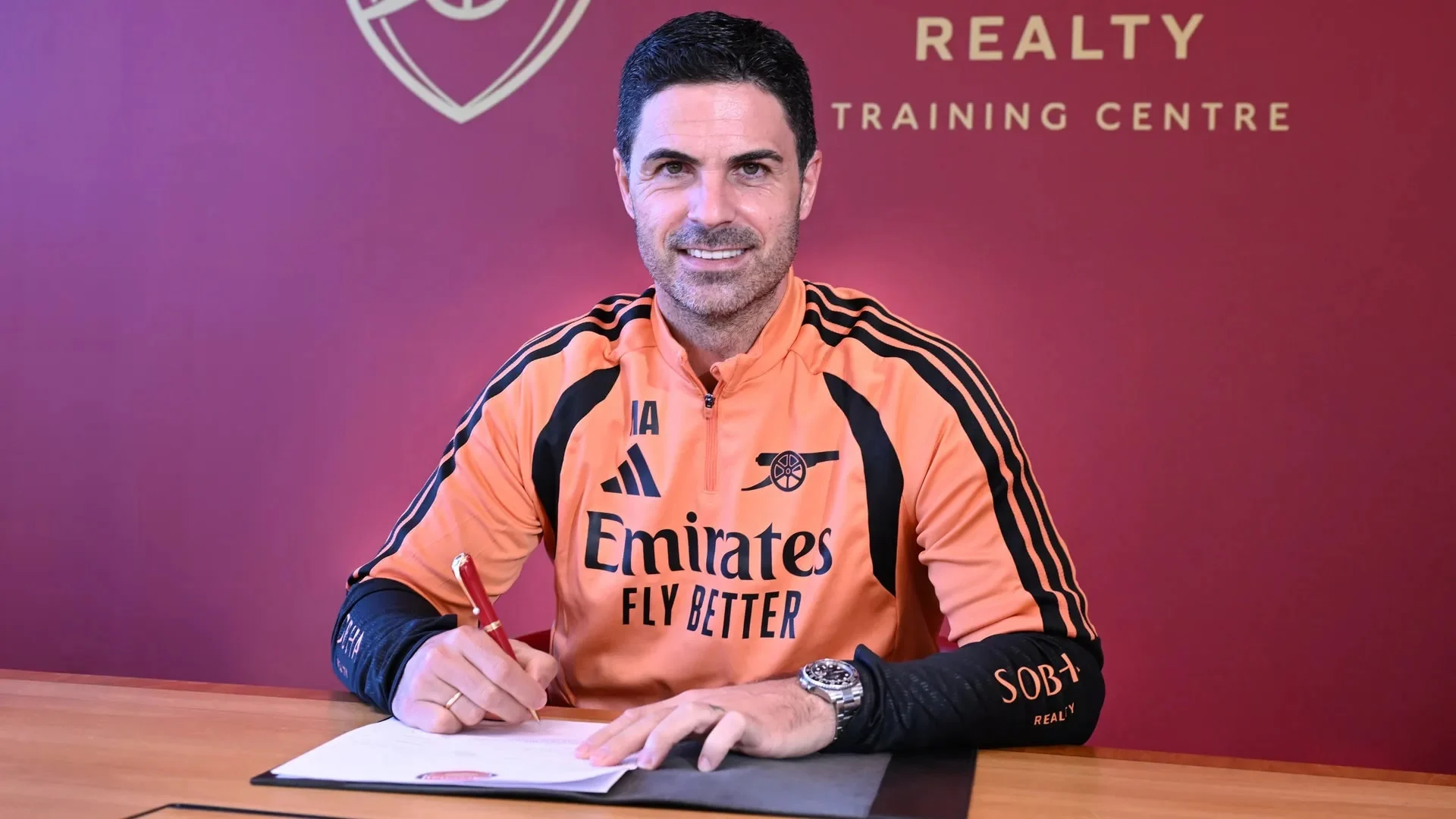 Mikel Arteta, in an orange Arsenal training top, smiles as he signs his new contract at the club's training centre