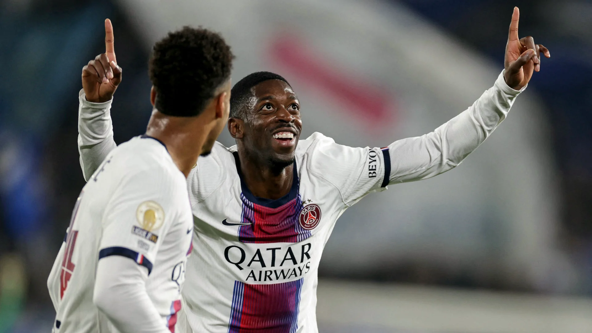 Ousmane Dembélé smiles and points both index fingers to the sky as he celebrates his goal against Strasbourg in Paris Saint-Germain's white away kit, with a team-mate in the foreground at the Stade de la Meinau. Photo: Romeo Boetzle/AFP via Getty Images (TSN)
