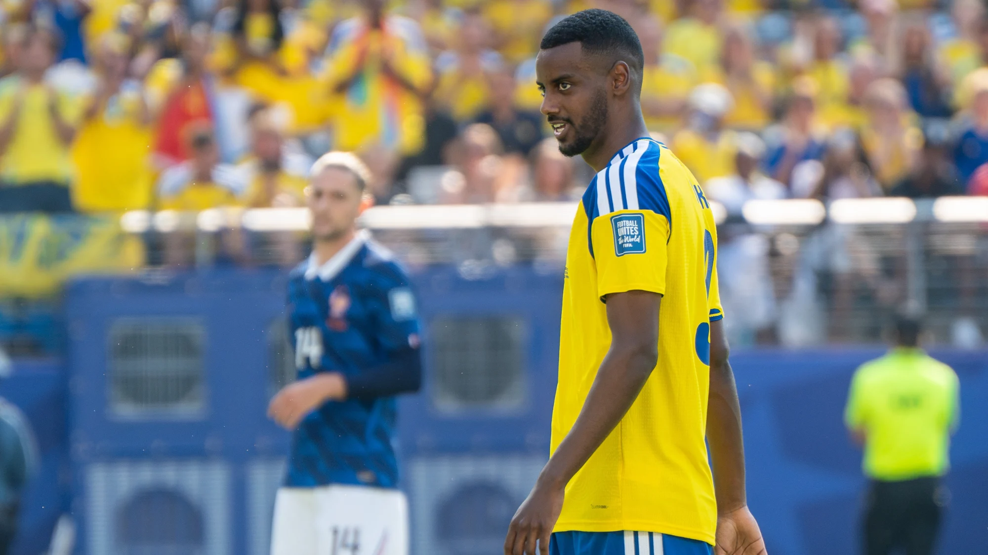 Alexander Isak in a yellow Sweden jersey looking over his shoulder during a match at a packed stadium.